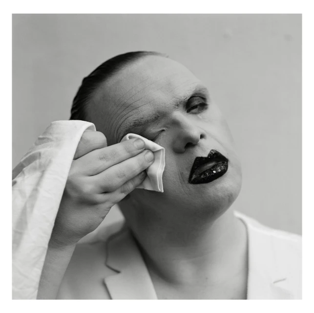 Close-up, black-and-white portrait of a person with heavy makeup wiping their eye with a white cloth, wearing a white jacket.
