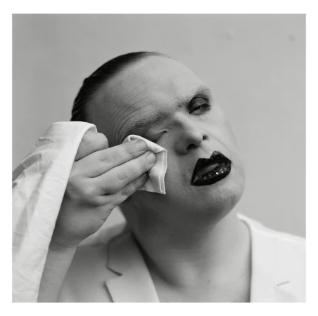 Close-up, black-and-white portrait of a person with heavy makeup wiping their eye with a white cloth, wearing a white jacket.