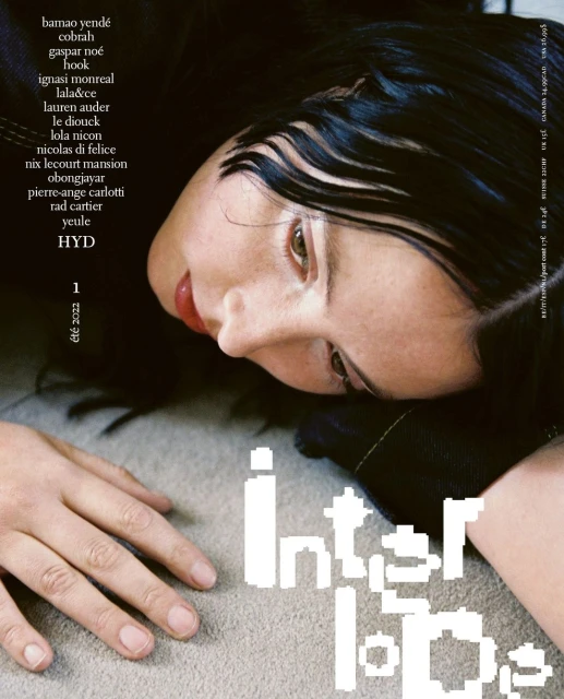 Fashion magazine cover featuring a young woman with black hair, lying on a neutral surface, and gazing downward in soft lighting.