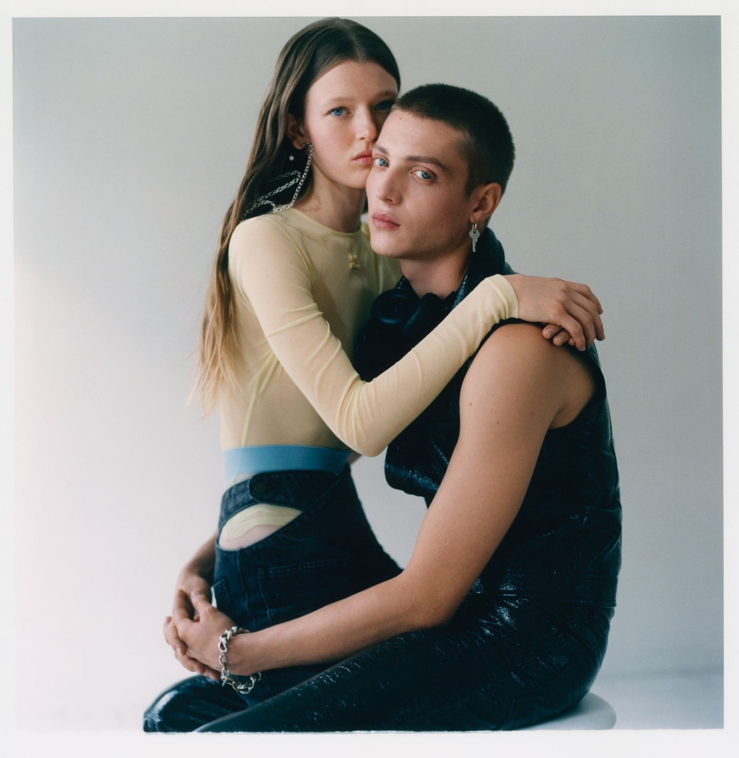 A young adult couple in a studio setting; the woman with long brown hair embraces the man wearing a glossy black vest.