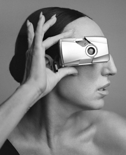 Monochrome portrait of a person holding a silver camera over one eye, with smooth hair, bare shoulder, and neutral expression.