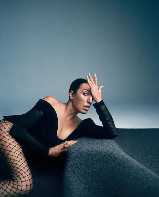 Adult woman in a black bodysuit and fishnet stockings poses in a studio on a gray surface with a gray to blue gradient background.