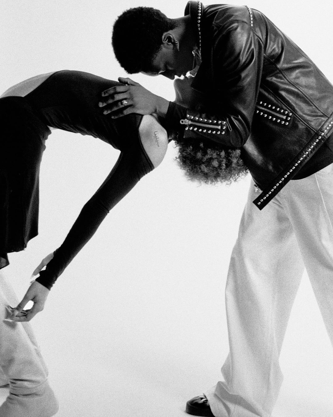Black and white image of a Black male wearing a studded leather jacket and white pants bending to touch heads with a Black female.