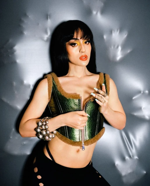 Woman in metallic green corset top with fur trim, posing against a crinkled silver background in a studio portrait with dramatic lighting.