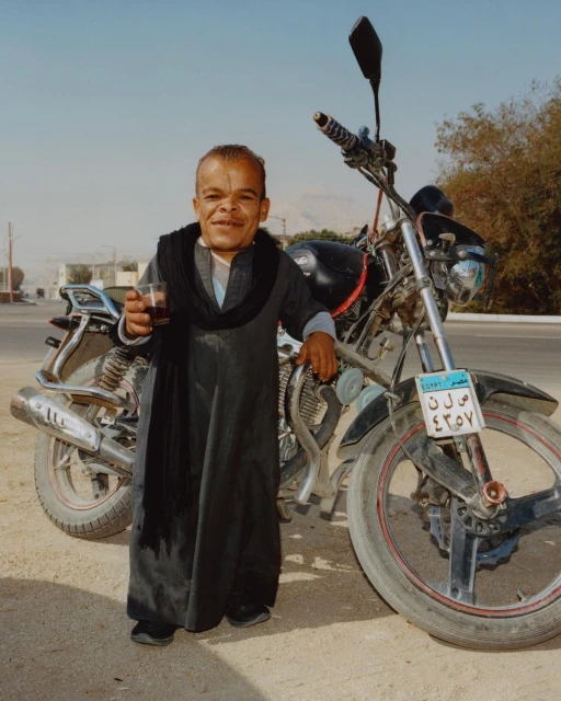 Adult male of small stature poses with a drink alongside a motorcycle on an outdoor road with buildings in the background under a clear sky.