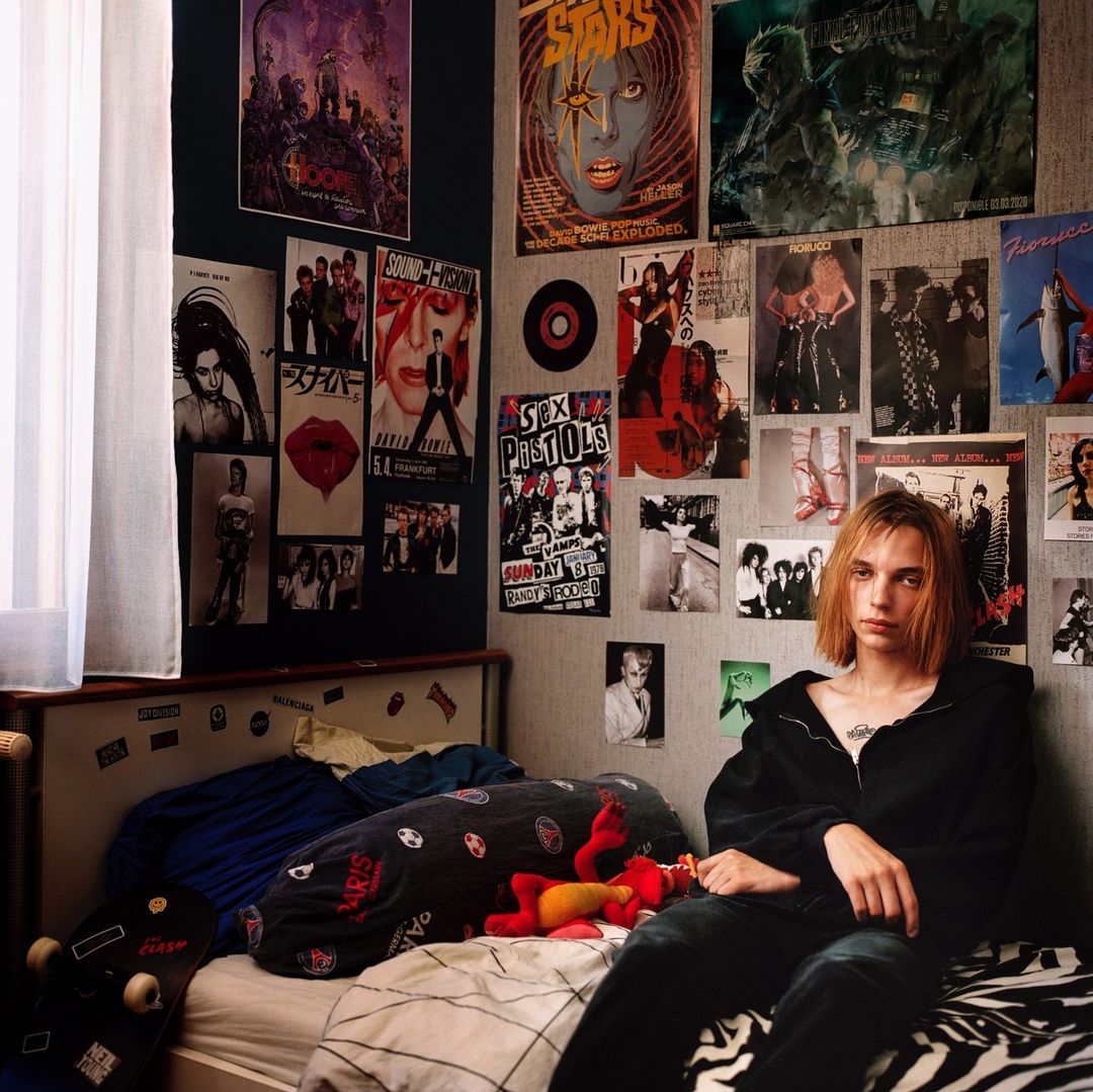 Teenager sits on a zebra-print bed in a bedroom decorated with posters. The room shows a young person's interest in music and pop culture.
