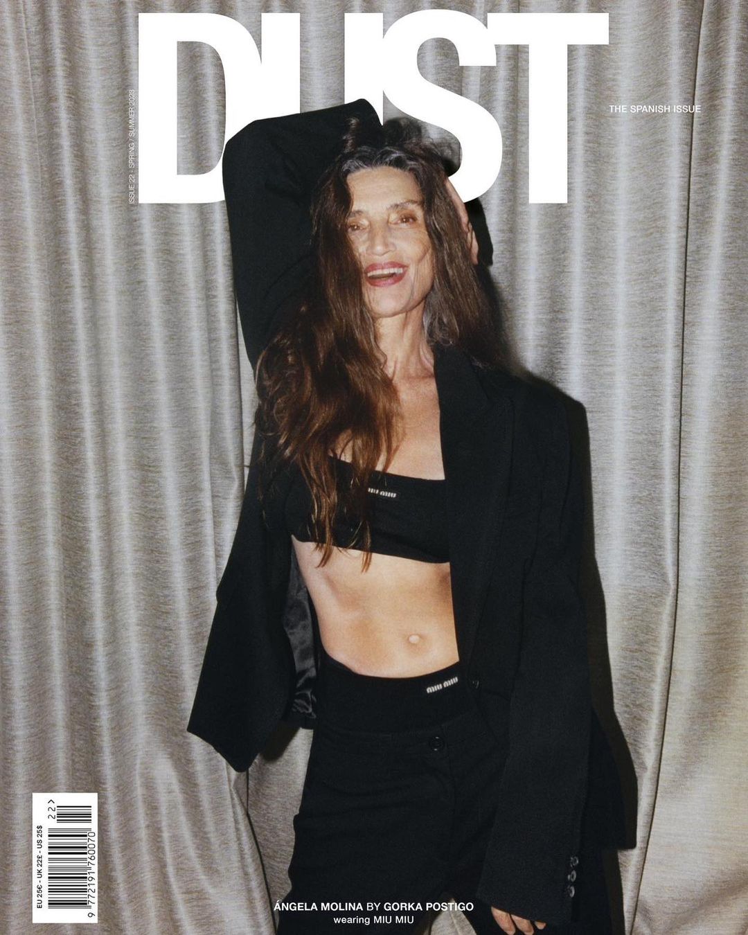 Magazine cover featuring an older woman with long brown hair wearing a black blazer and trousers against a textured curtain background.