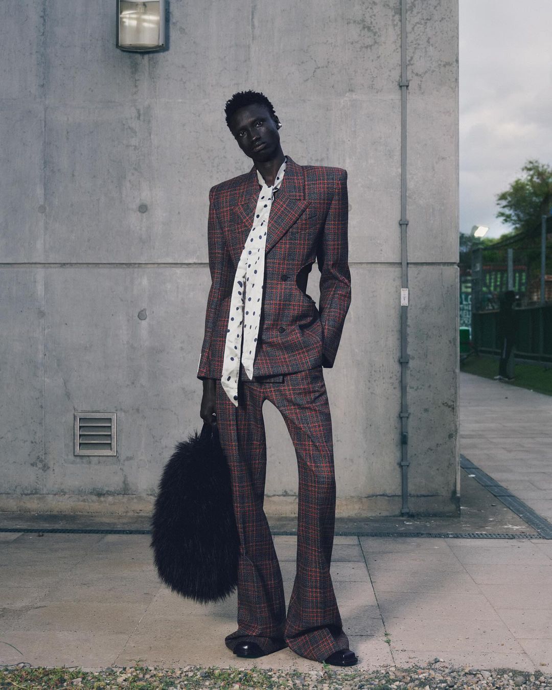 Fashionable adult male model wearing plaid suit, patterned tie, and holding a fur purse outdoors in an editorial fashion shoot.