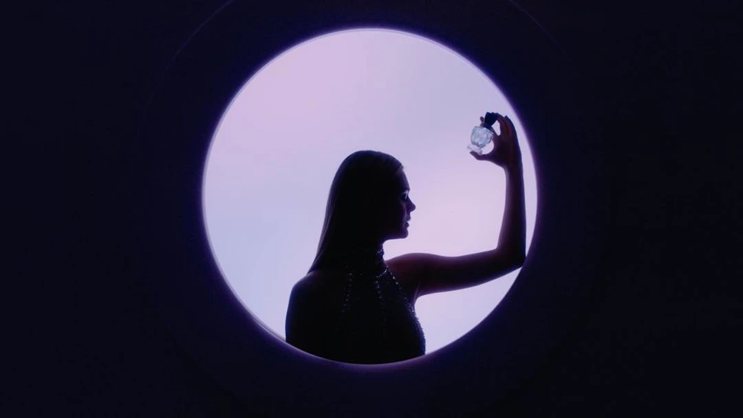 Silhouette of a woman in profile holding a clear perfume bottle, framed by a circular window against a graduated purple backdrop.