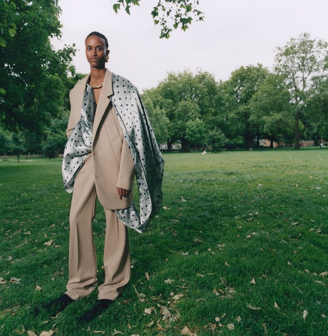 An adult stands in a park wearing a beige suit with a patterned scarf draped over the shoulder, showcasing fashion in a natural setting.