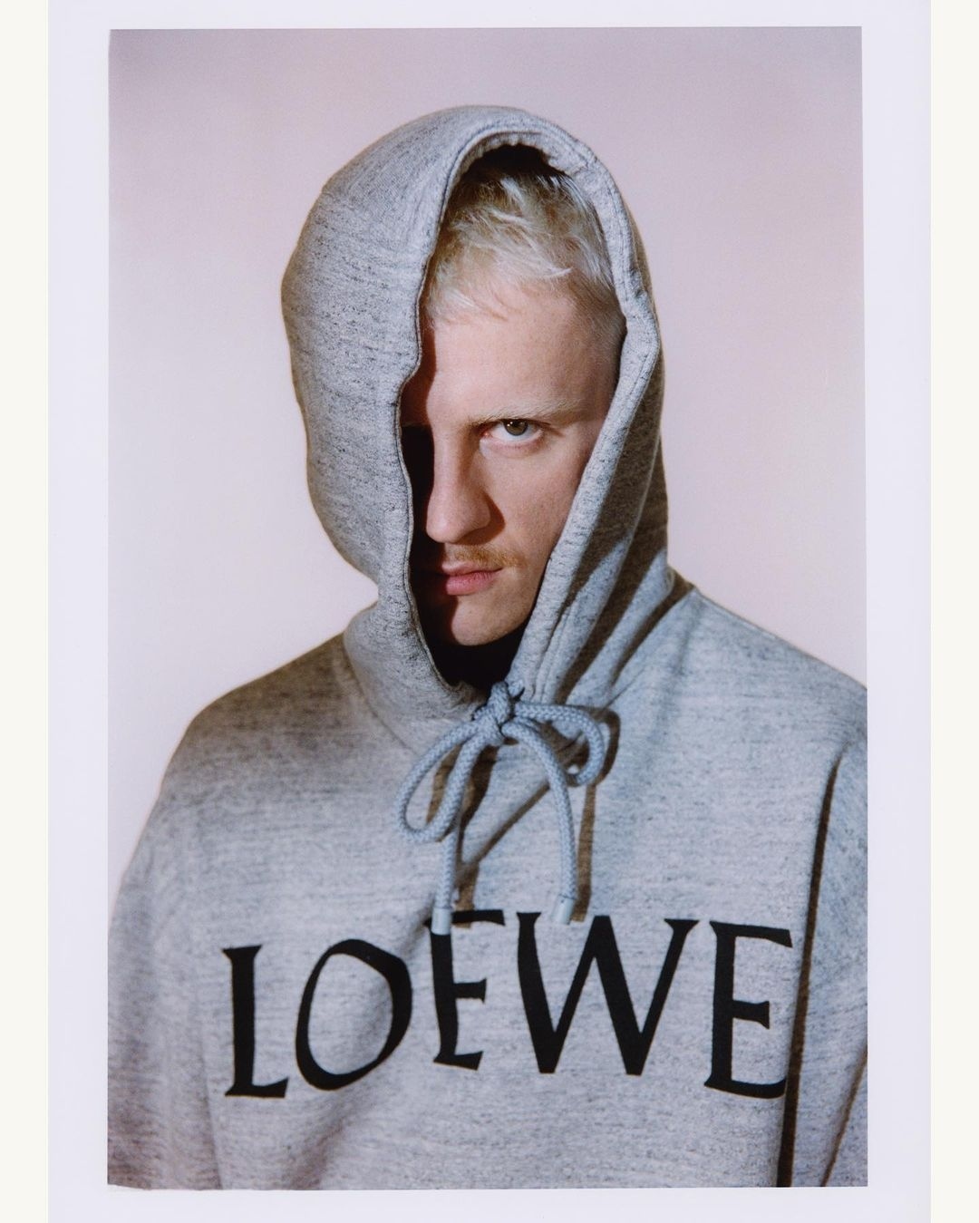 Portrait of a blond male model wearing a gray hooded sweatshirt with 'LOEWE' printed on the front, set against a neutral backdrop.