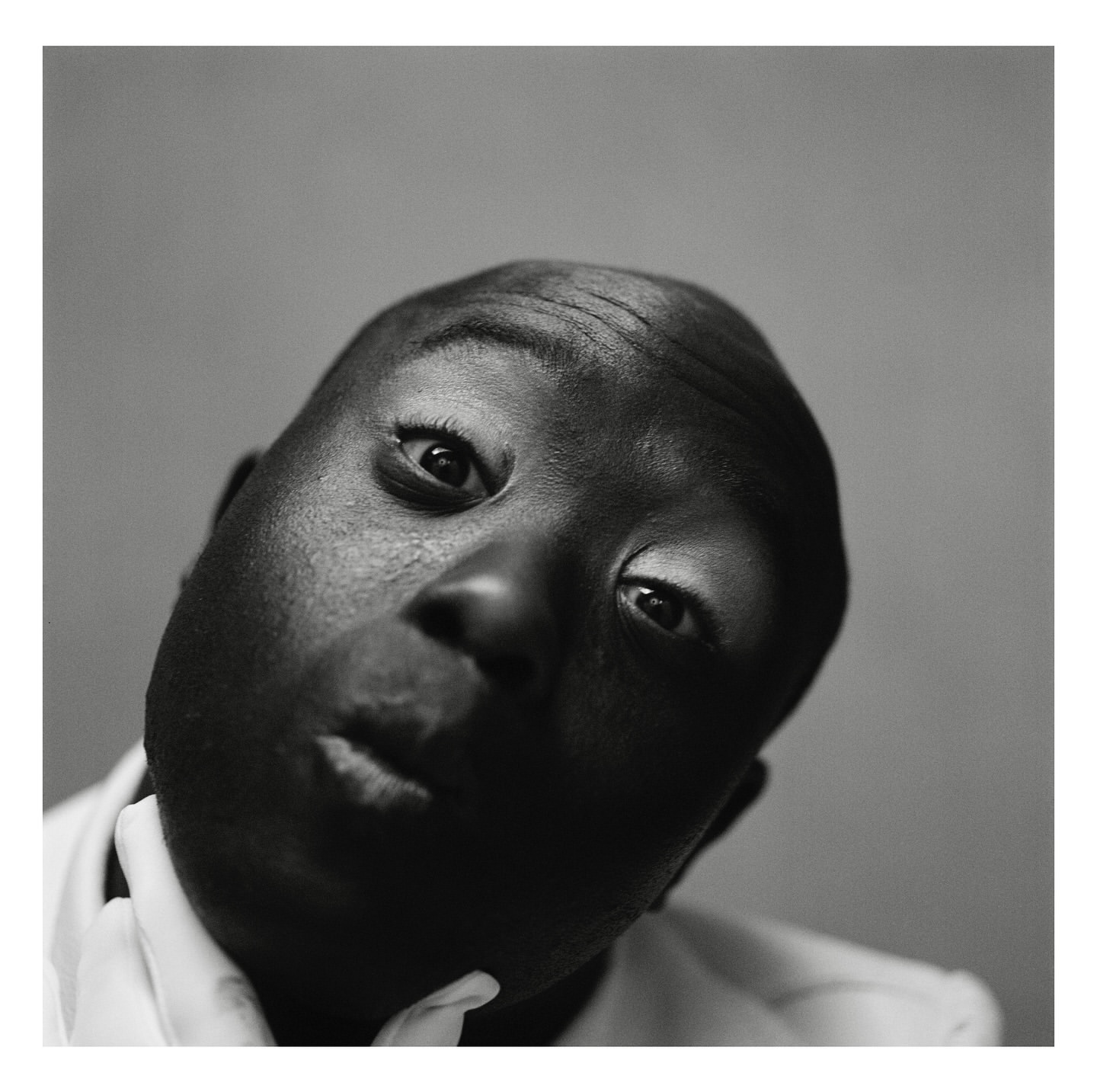 Close-up black and white portrait of an African American adult male looking up with a slight smirk and white shirt collar visible.