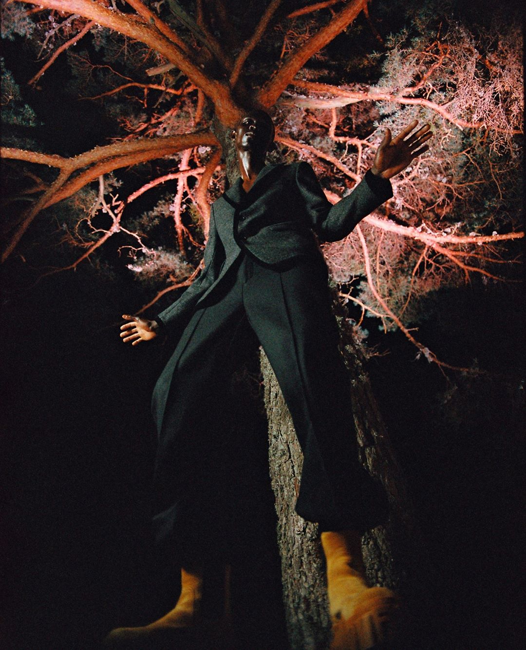 Model posing in a black suit and yellow boots in front of a tree with pink illuminated branches in a dimly lit outdoor setting.