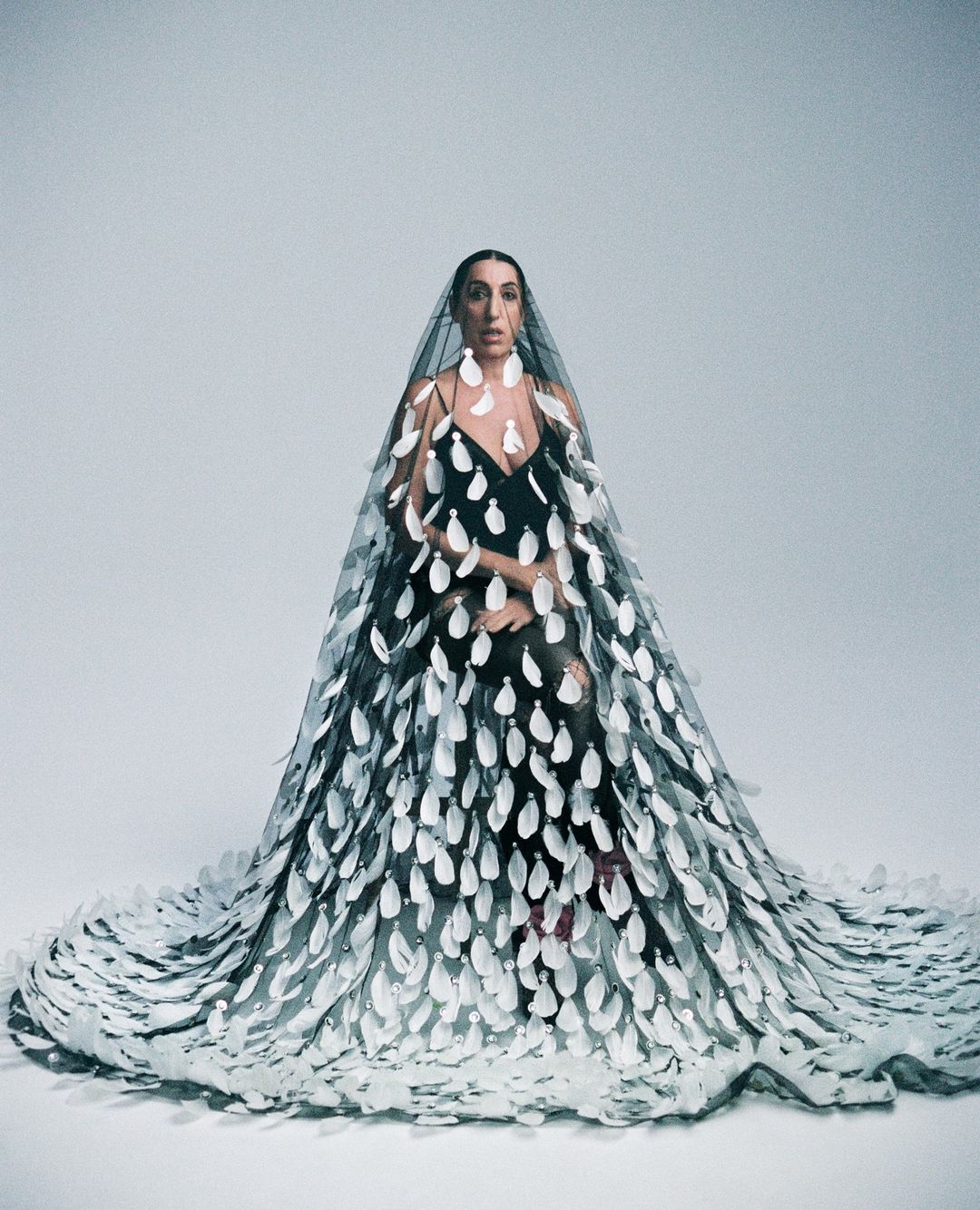 A woman poses in a black dress and sheer cape embellished with large white appliqués, set against a plain grey studio backdrop.