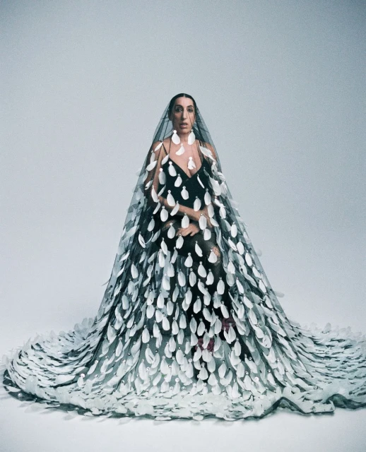 A woman poses in a black dress and sheer cape embellished with large white appliqués, set against a plain grey studio backdrop.