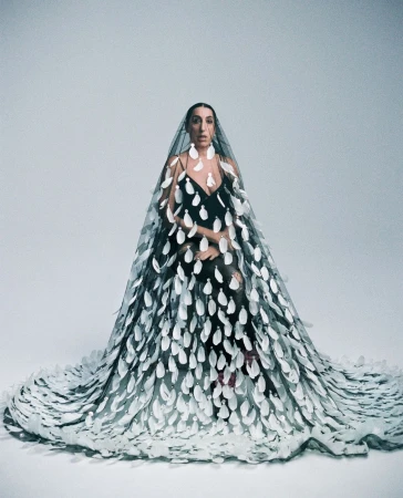 A woman poses in a black dress and sheer cape embellished with large white appliqués, set against a plain grey studio backdrop.