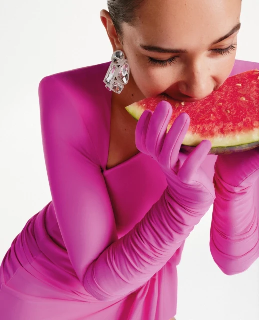 Woman in a pink dress and gloves bites into a slice of watermelon, posing in a studio with a white backdrop and wearing statement earrings.