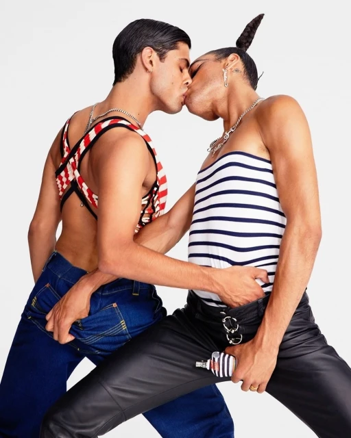 Two adult males kiss intimately. One wears a red and white top and blue jeans, while the other sports a striped top and leather pants in a studio shot.