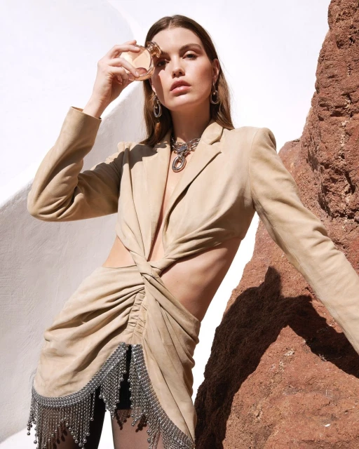 Woman in a beige cut-out dress and jewelry poses with perfume against a stark white wall and rock formation in an outdoor setting.