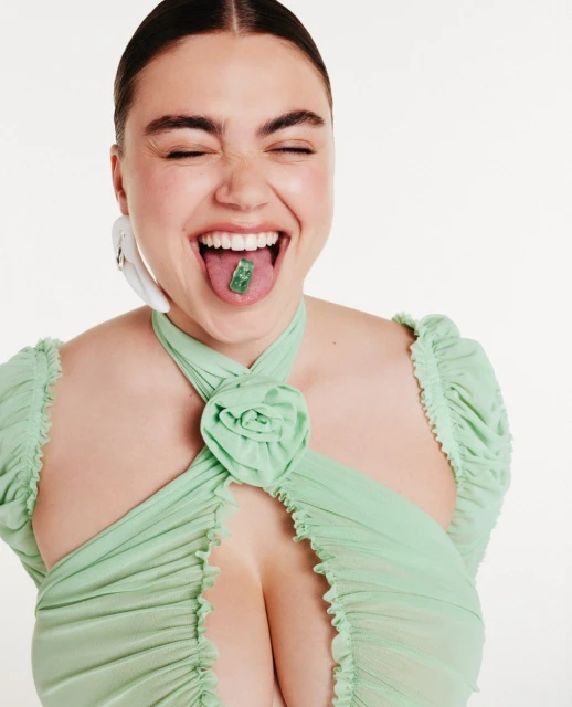 Smiling woman with a green gummy bear on her tongue, wearing a light green halter-style top, set against a bright white background.
