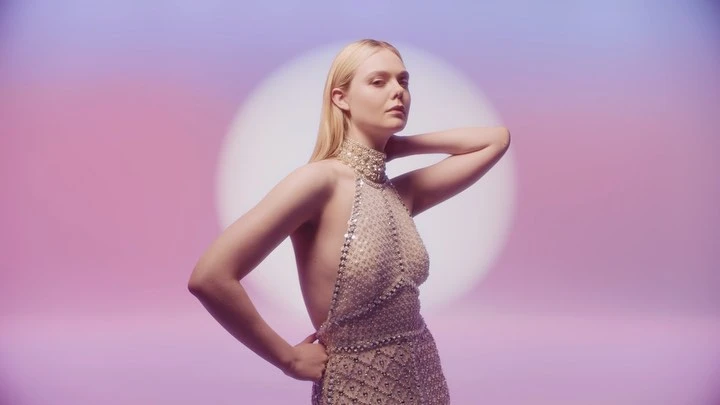 Glamorous portrait of a blonde woman wearing an elegant chainmail halter dress against a gradient pink and purple backdrop with a spotlight.