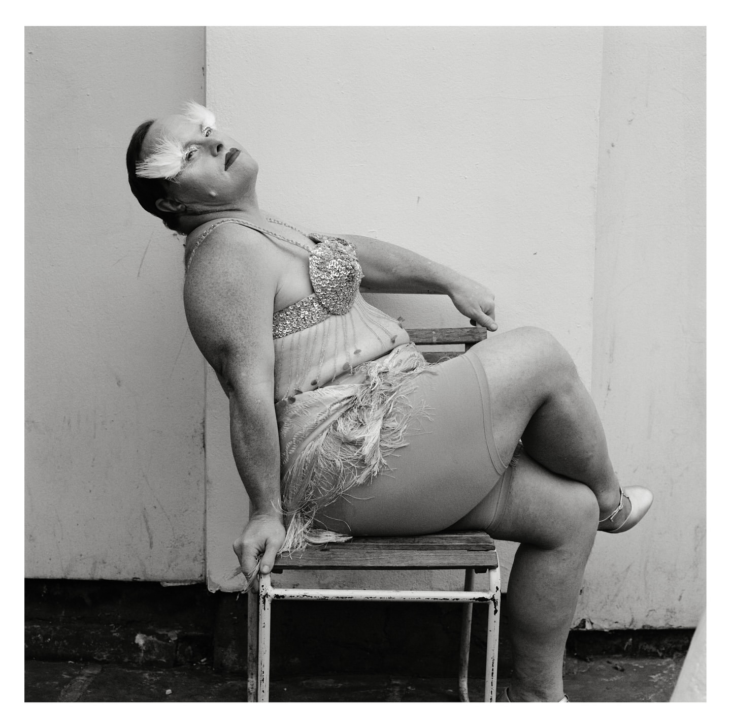 A vintage-style portrait of a mature adult in drag sitting on a chair outdoors, wearing a sequined dress and feathers, black and white.