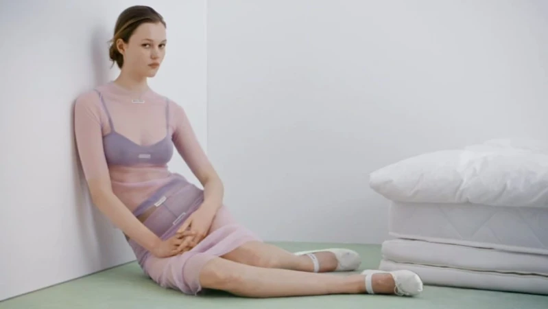 Adult female model in lavender sheer dress sits on the floor with a stack of mattresses in the background against a white wall.