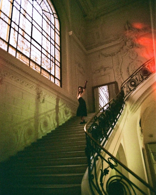 Woman poses on staircase with ornate railing beneath large arched window in a grand, vintage interior with green and red color grading.