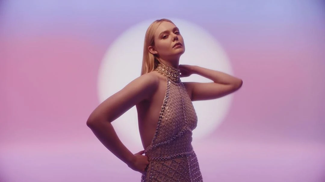 Blonde woman in jeweled halter dress poses in front of round bright light with pink and blue gradient background in medium shot.