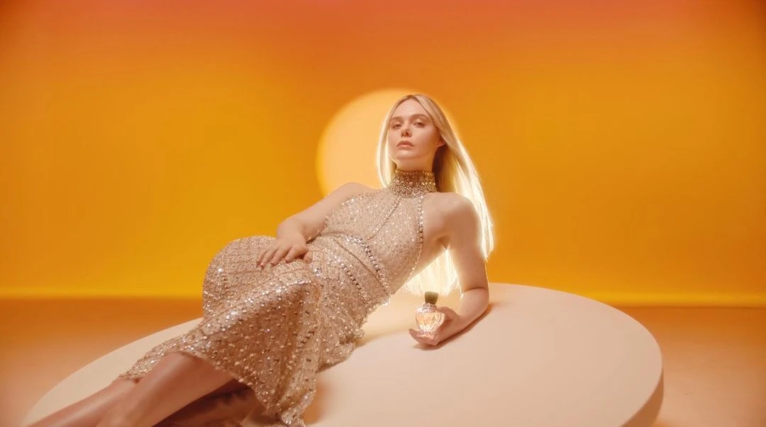 Elegant woman with blonde hair in a sparkling gown lies on a platform holding a perfume bottle, with a glowing orange background.