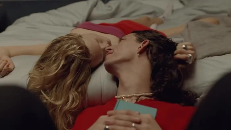 Two people lying down together on a bed, with a man holding a small book close to his chest while wearing rings and necklaces.