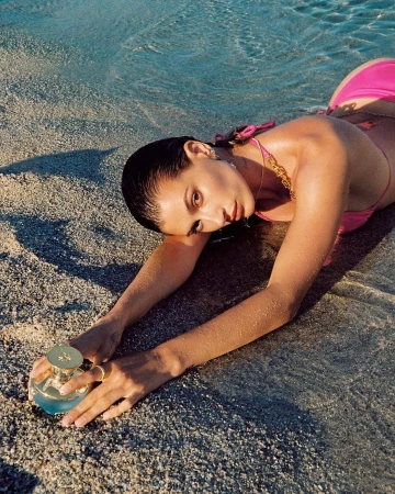 Woman in pink bikini lying on a sandy beach by shallow water, holding a round perfume bottle in her outstretched hands.