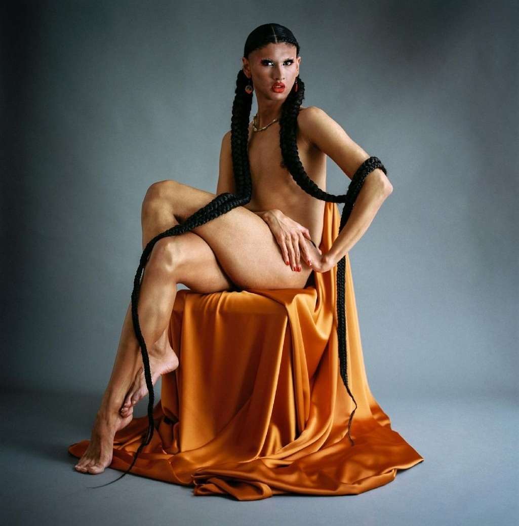 Unique portrait of a nude adult with long braids, orange jewelry, and red lips seated on an orange cloth against a grey backdrop.