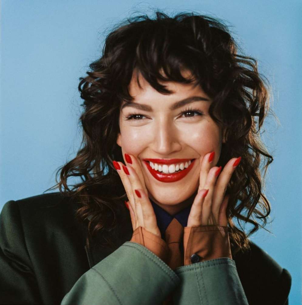 Close-up of smiling brunette woman with bright red lips and nails, wearing a dark green jacket against a light blue backdrop.