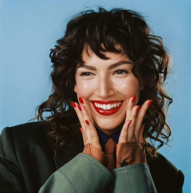 Close-up of smiling brunette woman with bright red lips and nails, wearing a dark green jacket against a light blue backdrop.