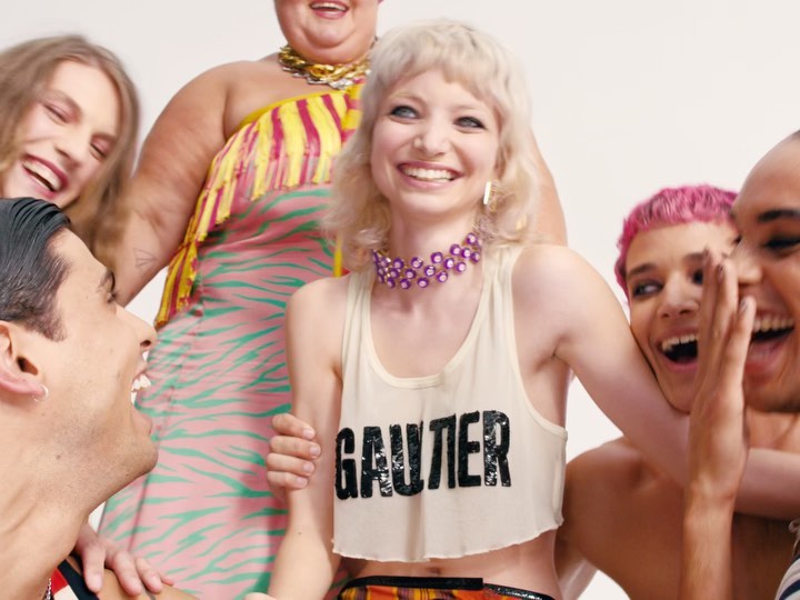 Group of six diverse young adults smiles in a brightly lit studio. A blonde woman in a white crop top is centered in the frame.