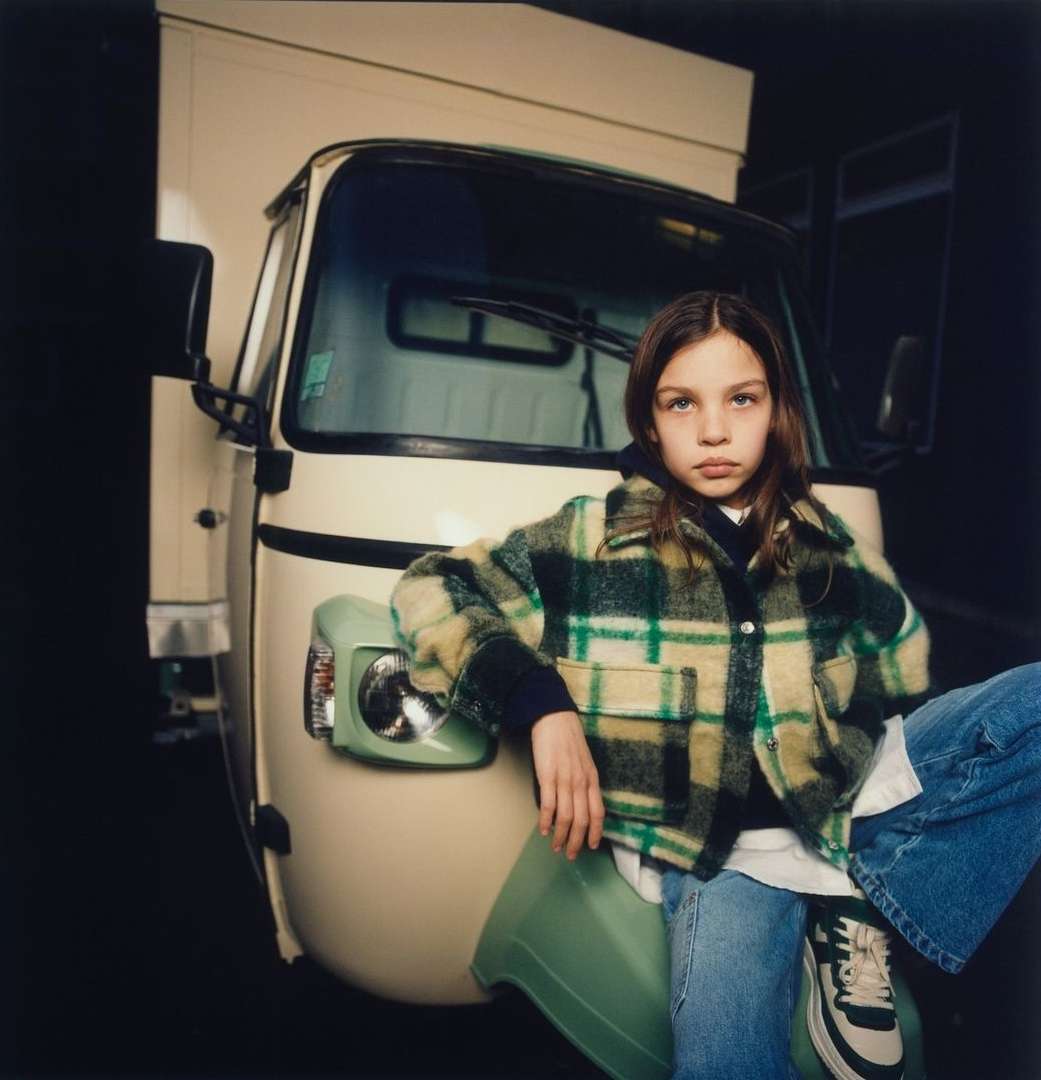 Young girl with straight brunette hair poses sitting on a green object in front of a cream-colored Piaggio Ape van.