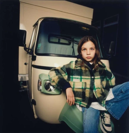 Young girl with straight brunette hair poses sitting on a green object in front of a cream-colored Piaggio Ape van.