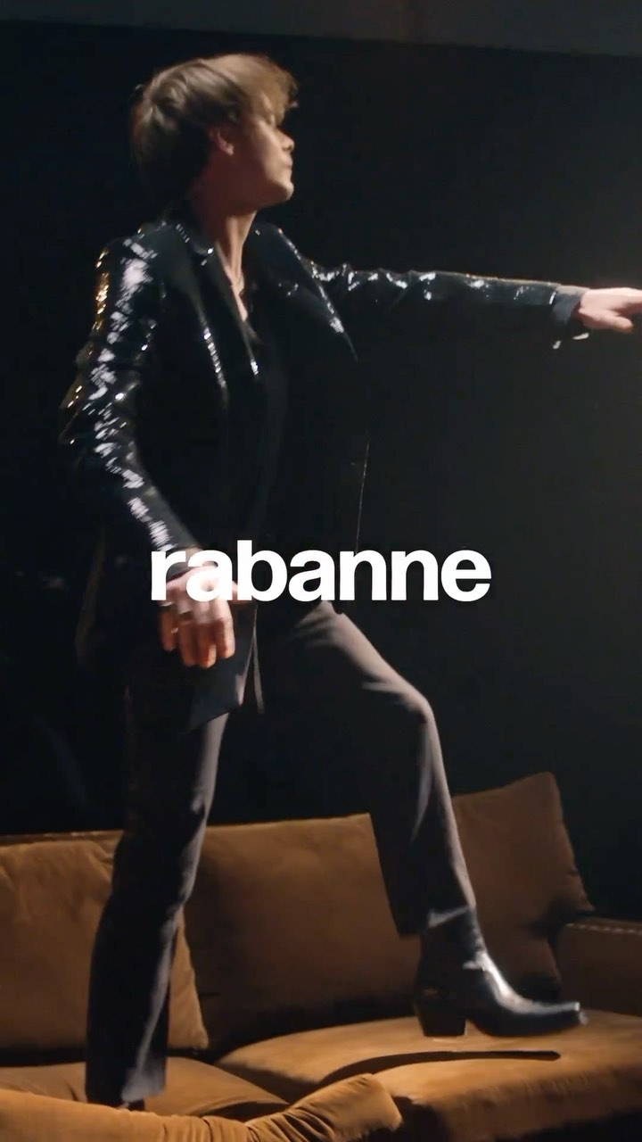 Fashion shot of a person wearing a black shiny blazer, trousers, and black boots, standing on a brown couch against a dark background.