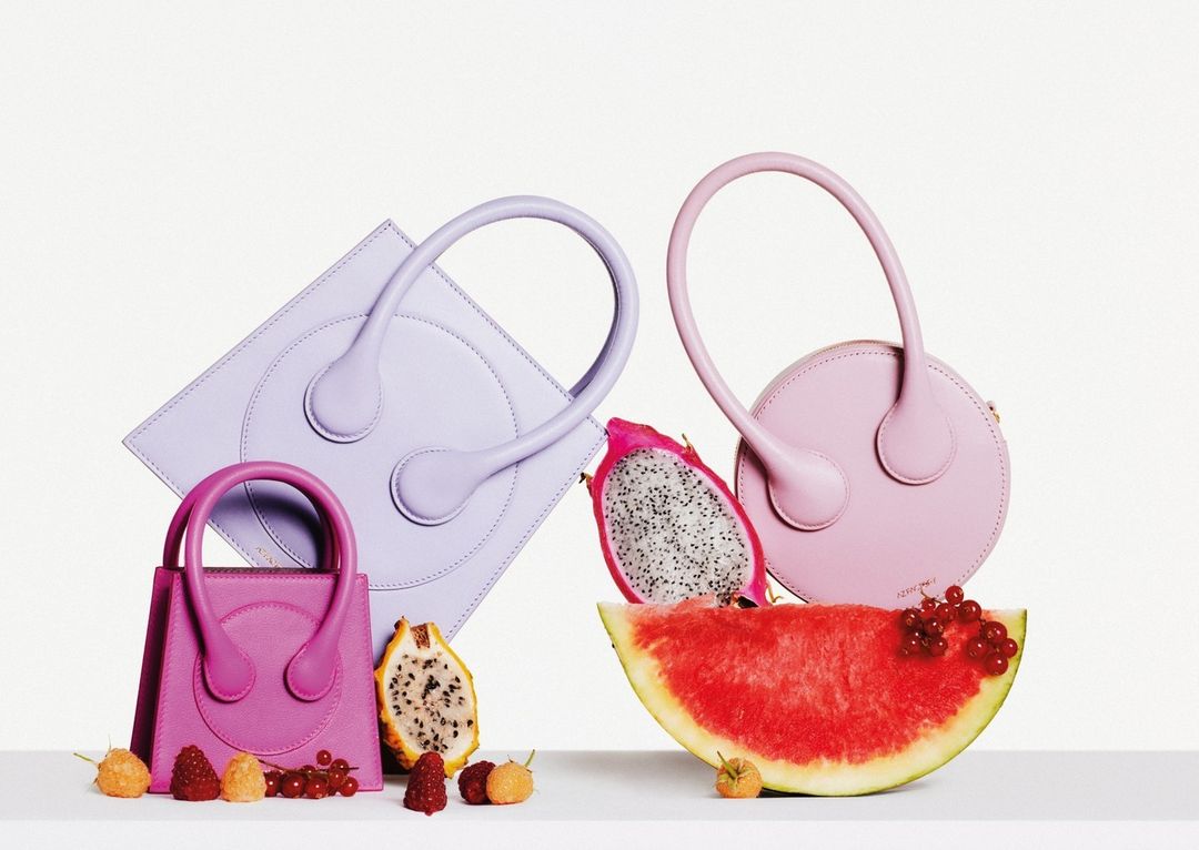 Three fashionable handbags in pink, purple, and magenta hues arranged with slices of watermelon and dragon fruit against a white background.