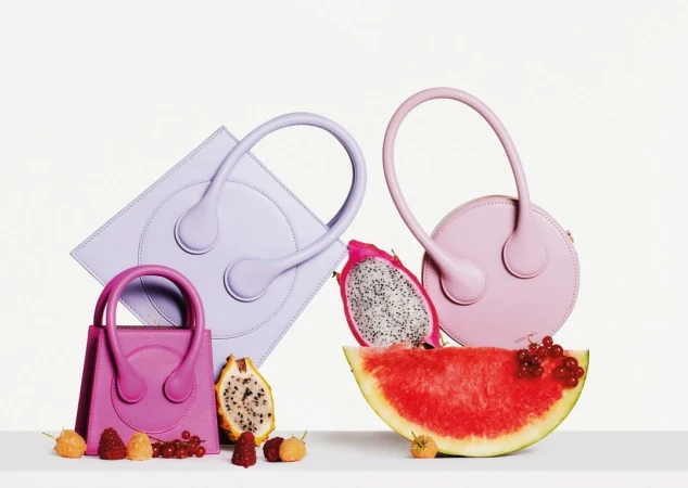 Three fashionable handbags in pink, purple, and magenta hues arranged with slices of watermelon and dragon fruit against a white background.