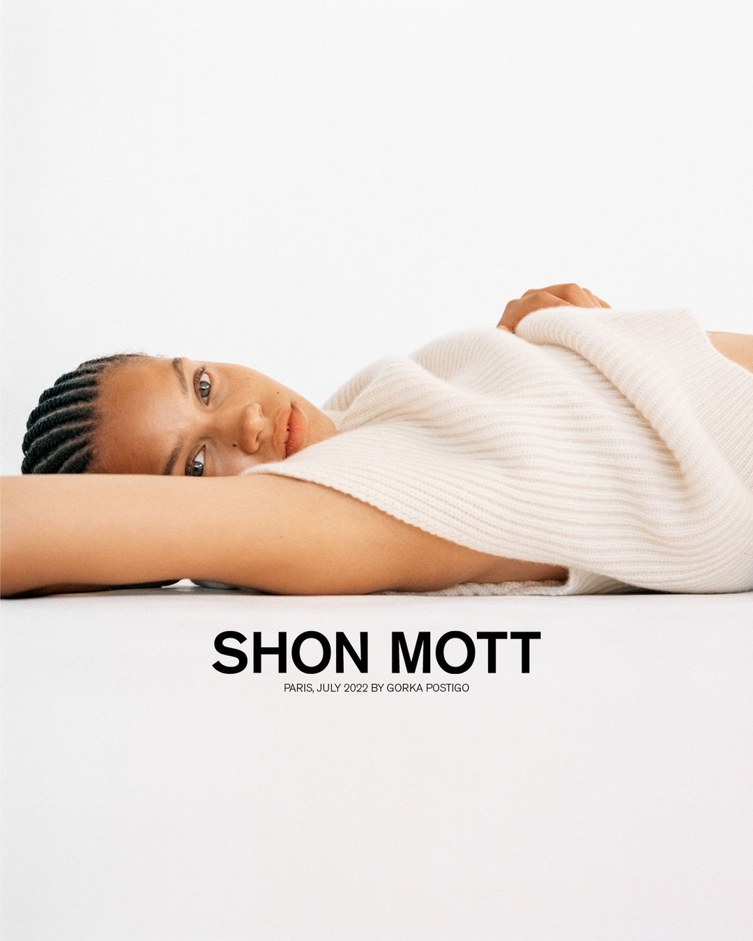 A woman with braided hair is lying down, wearing a cream-colored sweater, against a white background. Fashion photography with bright, soft lighting.