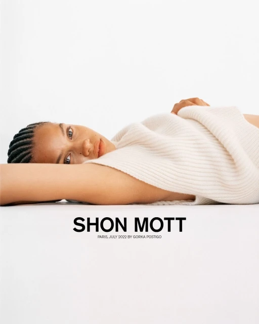 A woman with braided hair is lying down, wearing a cream-colored sweater, against a white background. Fashion photography with bright, soft lighting.