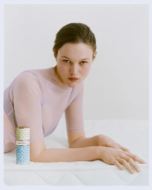 Fashion shot of a woman with light skin, wearing a lavender top, posed in front of a stack of cans in a light, airy environment.