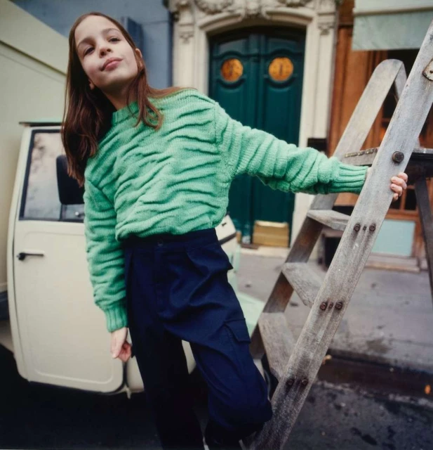 Young girl poses on a wooden ladder outdoors in a vibrant green patterned sweater and dark trousers, with a delivery van nearby.