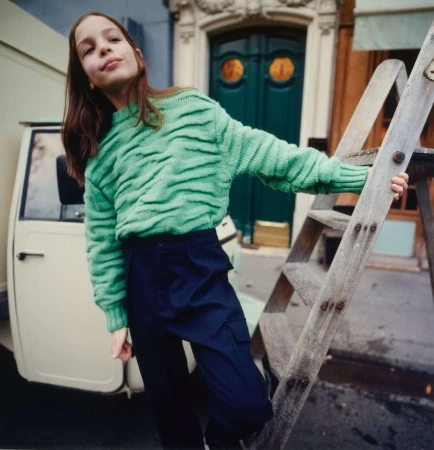 Young girl poses on a wooden ladder outdoors in a vibrant green patterned sweater and dark trousers, with a delivery van nearby.