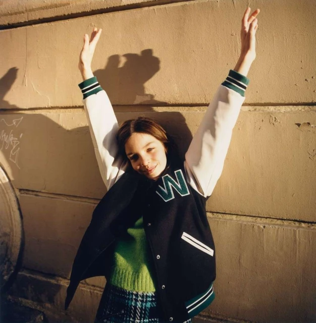 A young woman in a letterman jacket poses outdoors with her arms raised, creating shadows on the wall behind her.