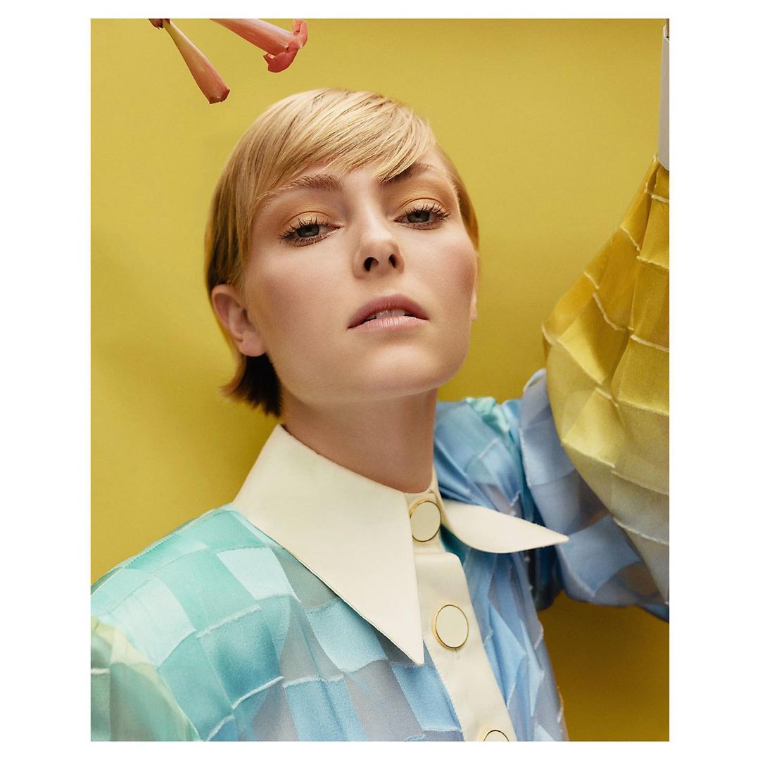Close-up portrait of a woman with short blonde hair, wearing a color block shirt with a yellow background and pink flowers above.