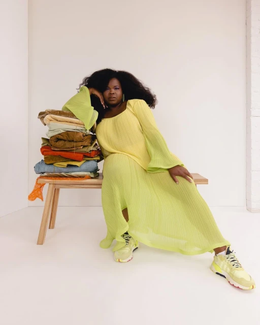 Elegant African-American woman in a lime green dress poses next to a stack of colorful clothing on a wooden bench indoors.