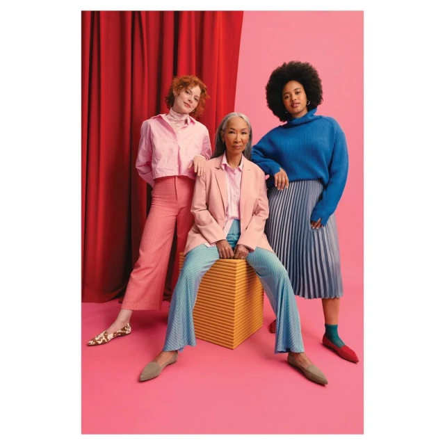 Three women of diverse ages pose in a colorful studio setting, wearing pink and blue attire with a pink and red backdrop.